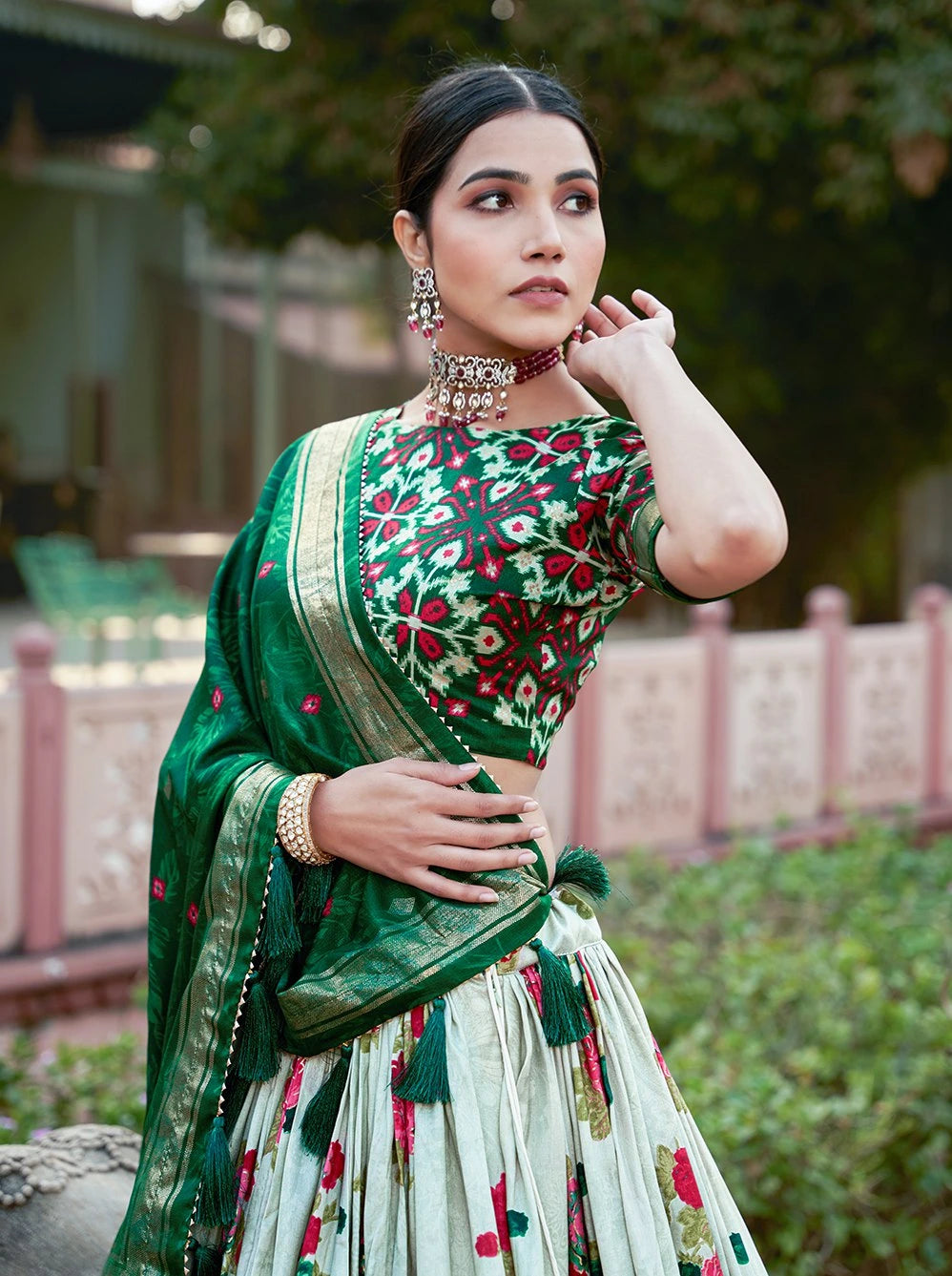 Woman wearing a green and red traditional outfit with jewelry, standing outdoors.